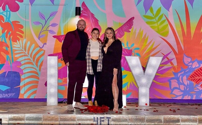 One Moment Hawaii photo of a man, woman, and teenager with their arms around each other, facing the camera in front of a colorful, tropical pink mural with marquis lettering as a backdrop.