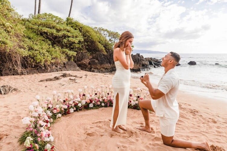 Engaged on Maui proposal planning photo: a man is down on one knee proposing to his girlfriend on the beach, surrounded by an array of flowers.